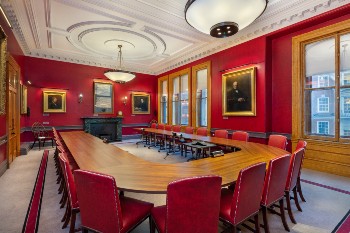 council meeting room with huge sash windows, red walls with various portraits hung on them, a fireplace and a u shaped table with red seats around it